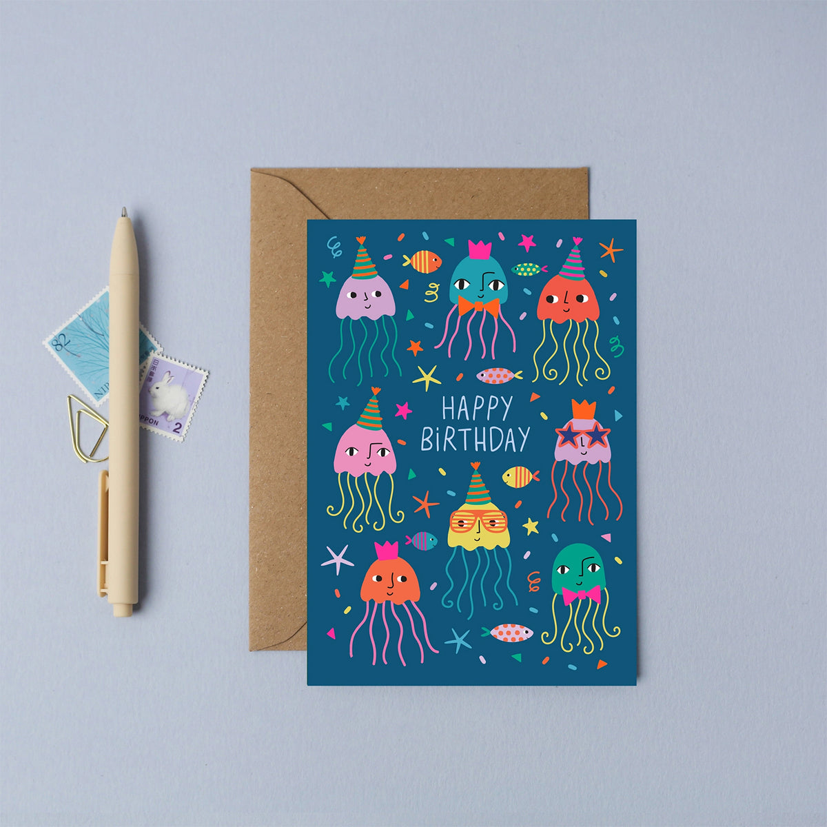 A birthday card with a brown envelope, styled on a light blue-gray surface next to a pen and two small postage stamps. The card features a dark blue underwater scene with colourful cartoon jellyfish, fish, and the words 'HAPPY BIRTHDAY'.