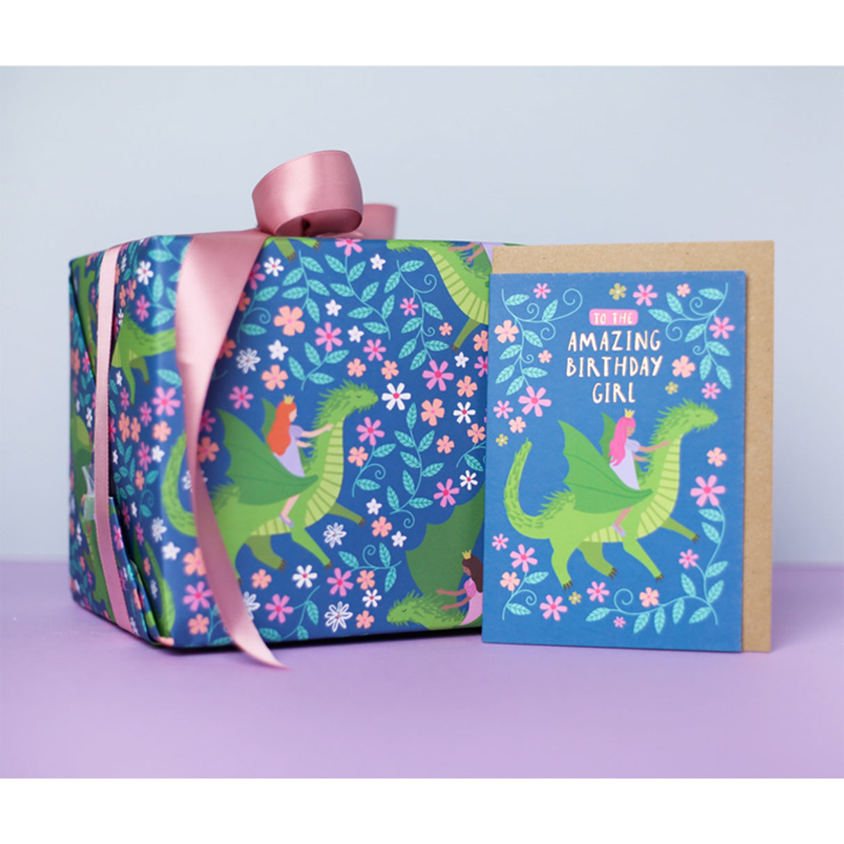 A wrapped gift box and matching greeting card featuring a colourful dragon and floral pattern. The box is wrapped in blue wrapping paper with green dragons, pink flowers, and a pink ribbon.