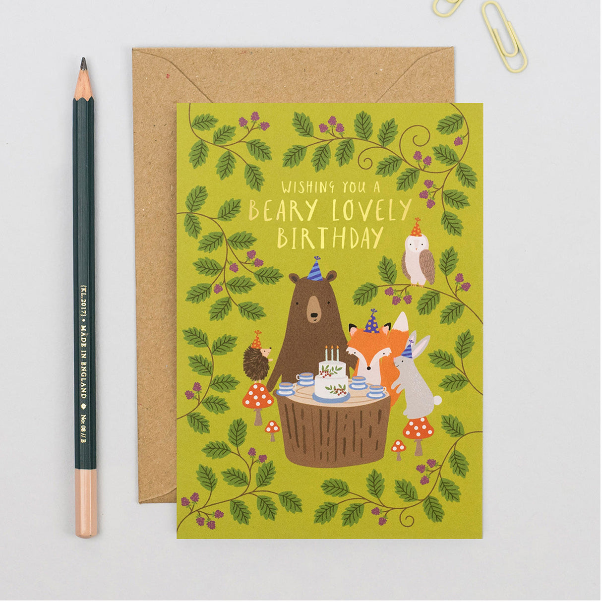 Birthday card with cartoon bears and foxes around a cake, next to a pencil on a light gray background