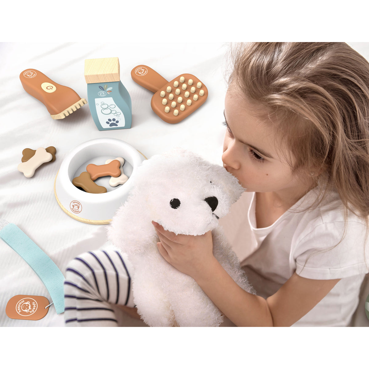 A young girl holding a white dog soft toy. Surrounding her are wooden pet care toys, including a brush, a foot, a milk carton, and a bowl with wooden bones.