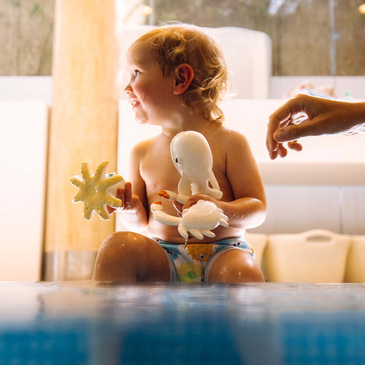 Child playing with bath toys in a bathtub, with a hand reaching in from the right side.