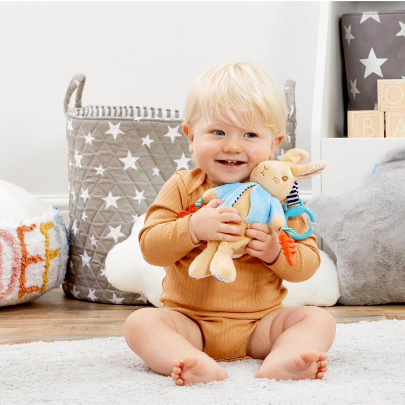 Baby holding a plush Peter Rabbut baby toy in a nursery setting with toys and a basket in the background.