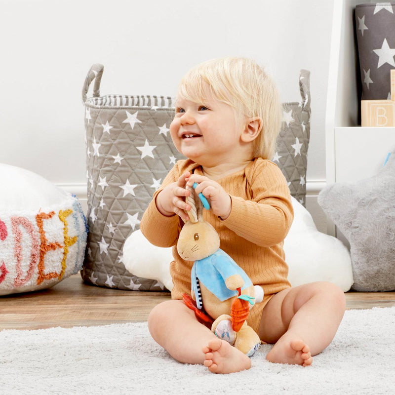 Baby holding a plush Peter Rabbut baby toy in a nursery setting with toys and a basket in the background.