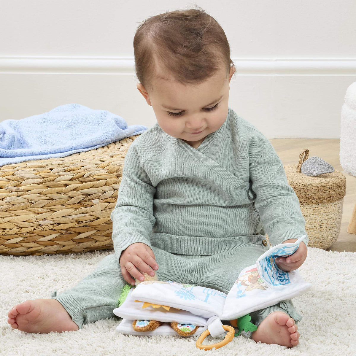 Baby sitting on a cream rug exploring the Peter Rabbit fold out activity book, with sensory tabs and crinkle pages