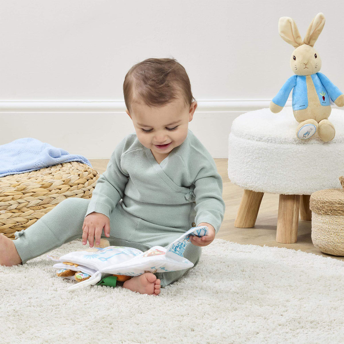 Smiling baby on a cream rug holding the Peter Rabbit fold out activity book, with a Peter Rabbit soft toy on a stool in the background