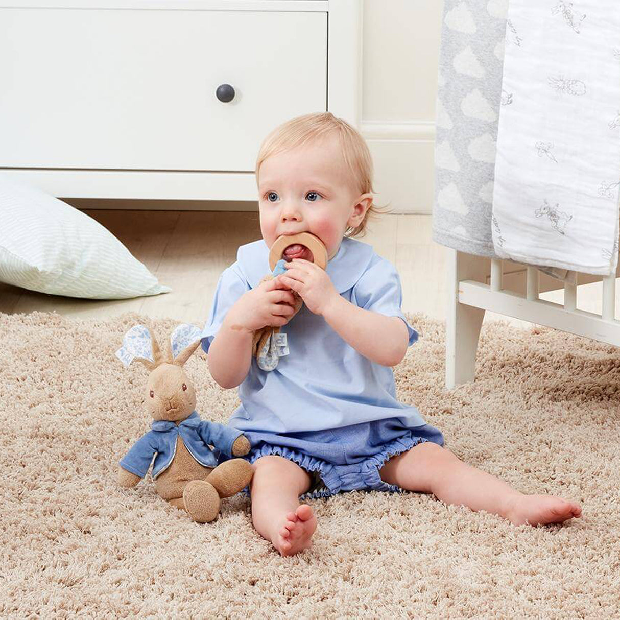 Baby sitting on a carpeted floor holding a wooden toy, with a teddy bear beside them.