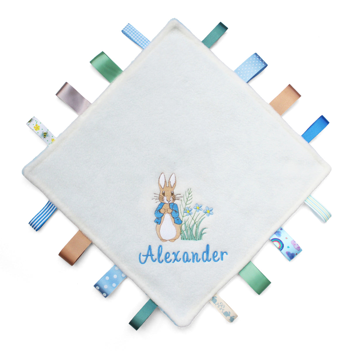 A personalised baby taggy blanket featuring an embroidered illustration of Peter Rabbit and the name 'Alexander' in blue script. The corners of the square blanket are finished with tabs of various colorful patterned ribbons, including blue, green, and tan.