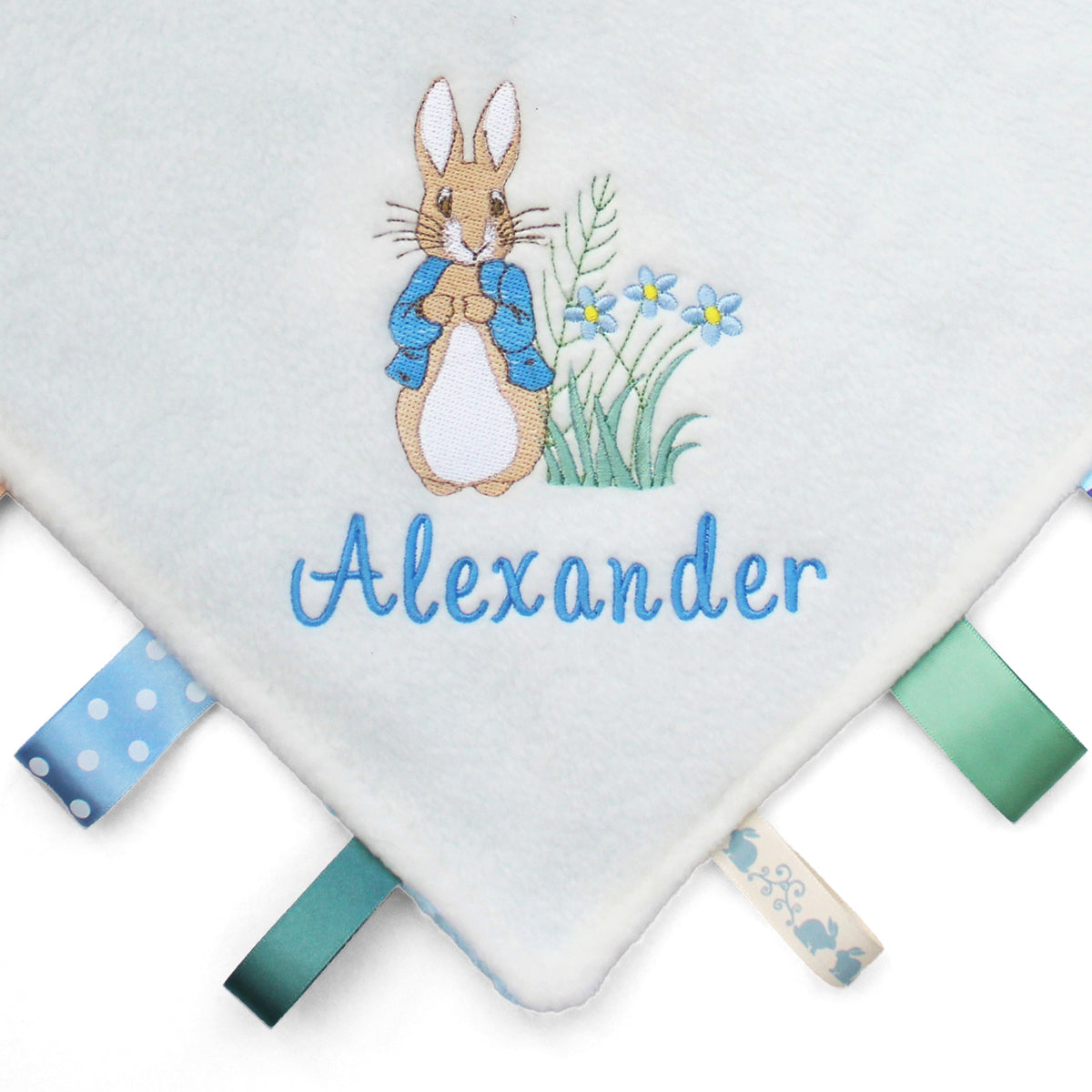 A close-up of a baby taggy blanket featuring an embroidered Peter Rabbit design, the name 'Alexander' in blue script, and colourful ribbon tabs along the edges.