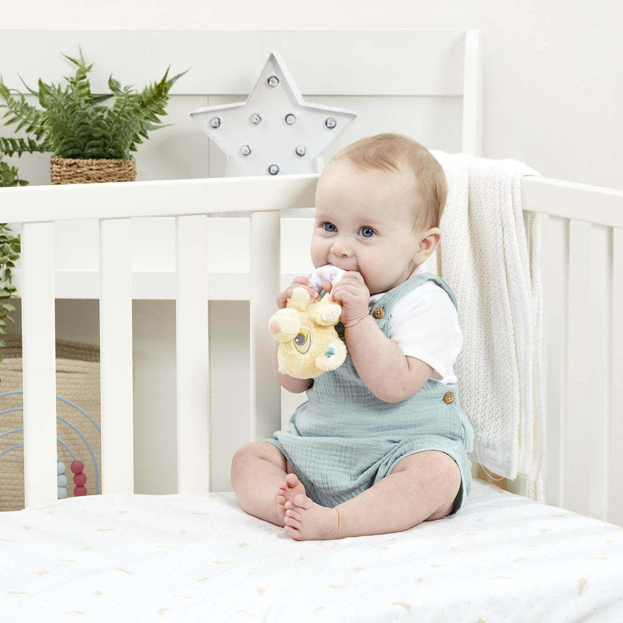 Baby sitting in a crib holding a Disney Lion King ring rattle with a white crib and decorative elements in the background.