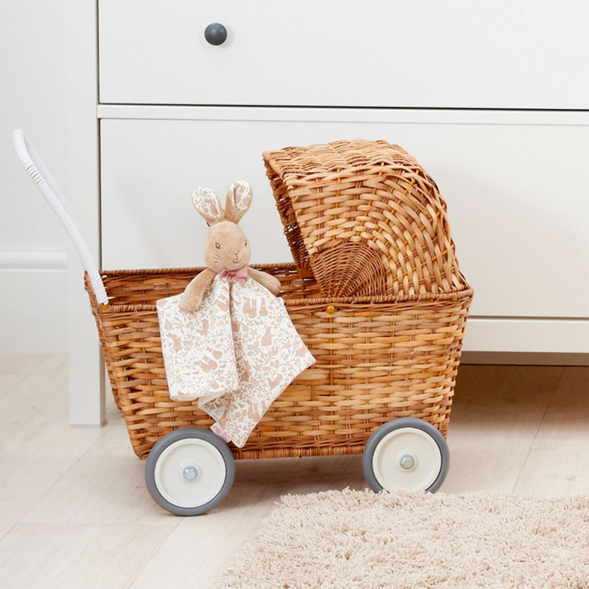 Wicker baby buggy with plush flopsy bunny toy blanket on a light wooden floor.