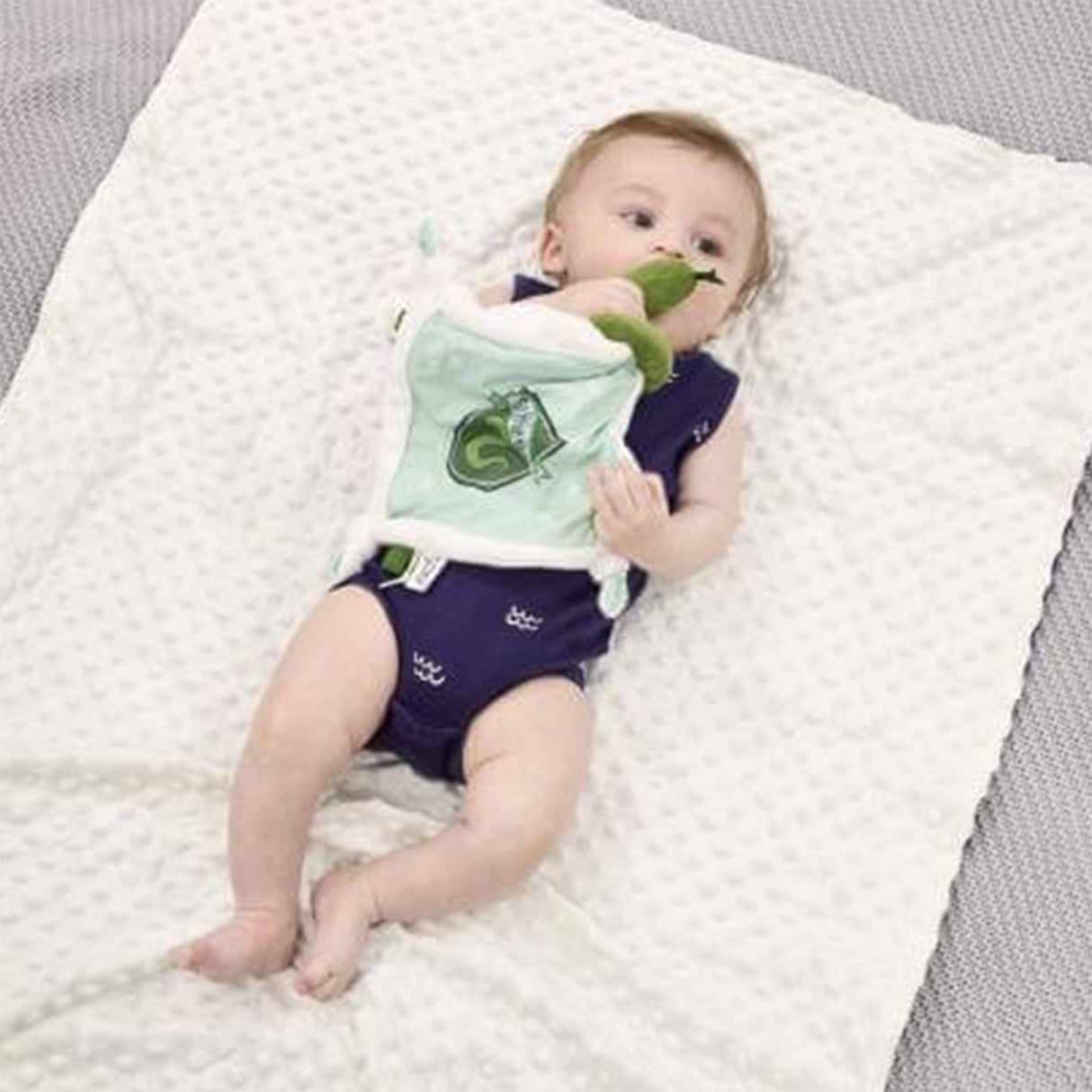 Baby lying on a textured surface with a plush Slytherin comfort blanket with a green and white color scheme, featuring a snake design, Slytherin crest, and embroidered stars.