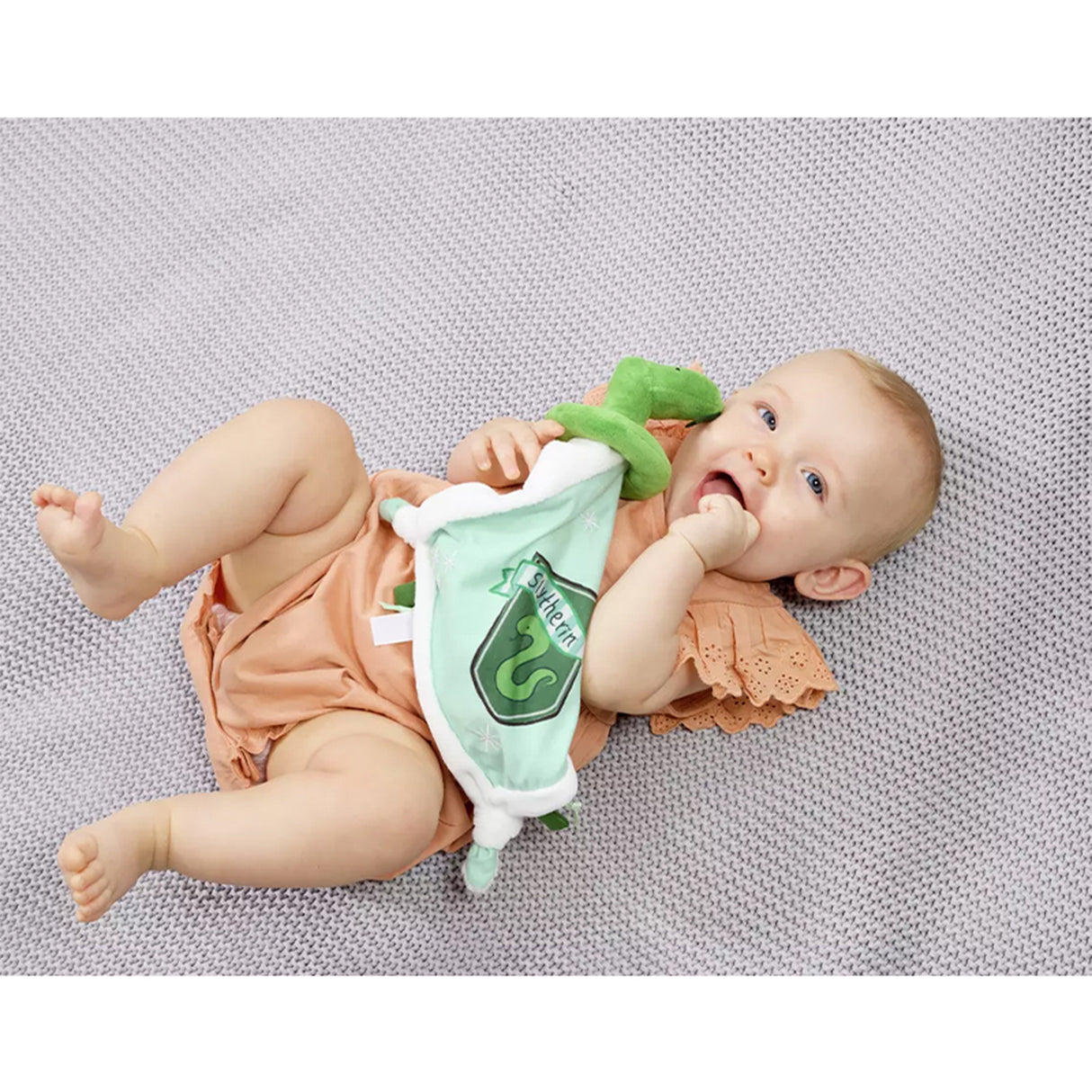 Baby lying on a textured surface with a plush Slytherin comfort blanket with a green and white color scheme, featuring a snake design, Slytherin crest, and embroidered stars.