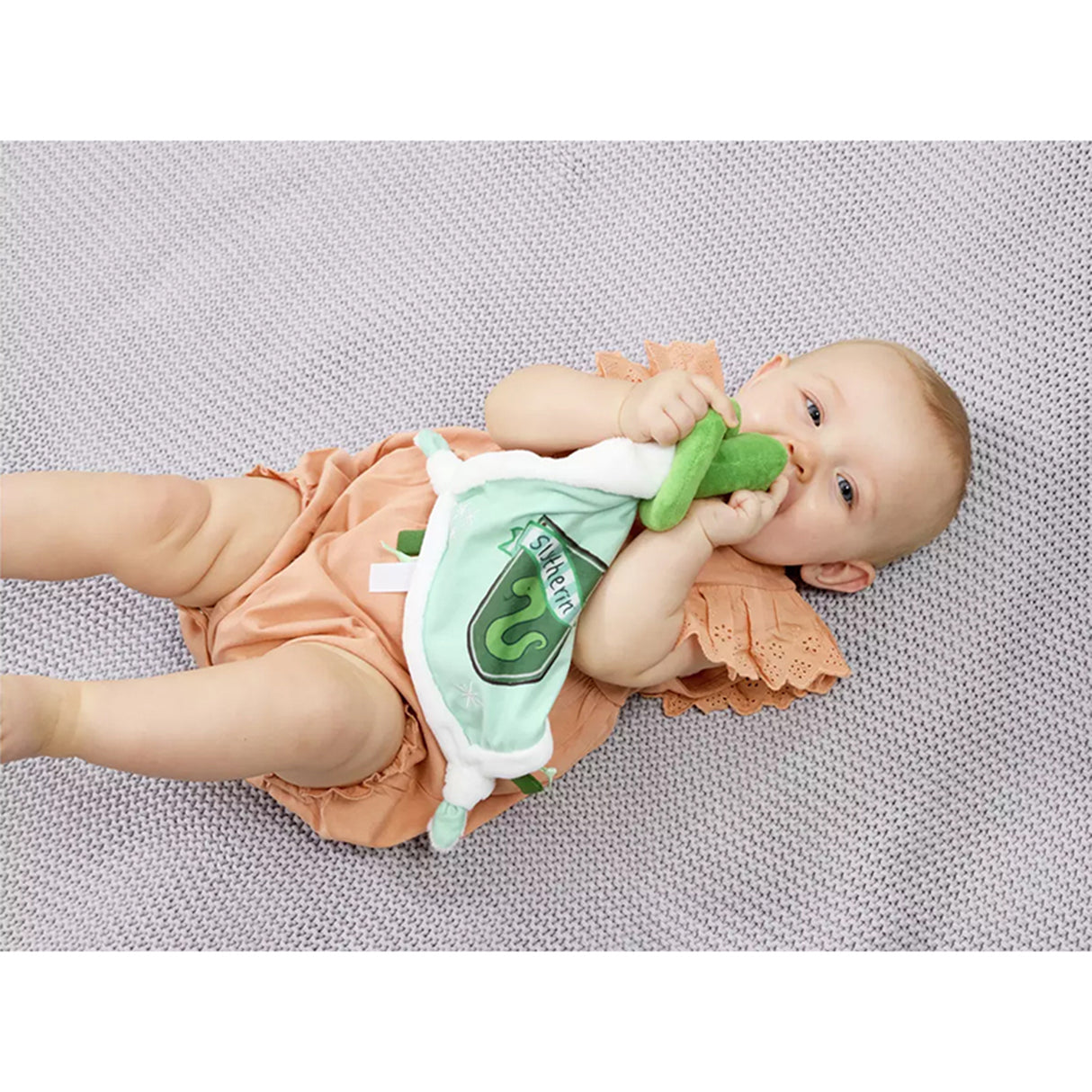 Baby lying on a textured surface with a plush Slytherin comfort blanket with a green and white color scheme, featuring a snake design, Slytherin crest, and embroidered stars.
