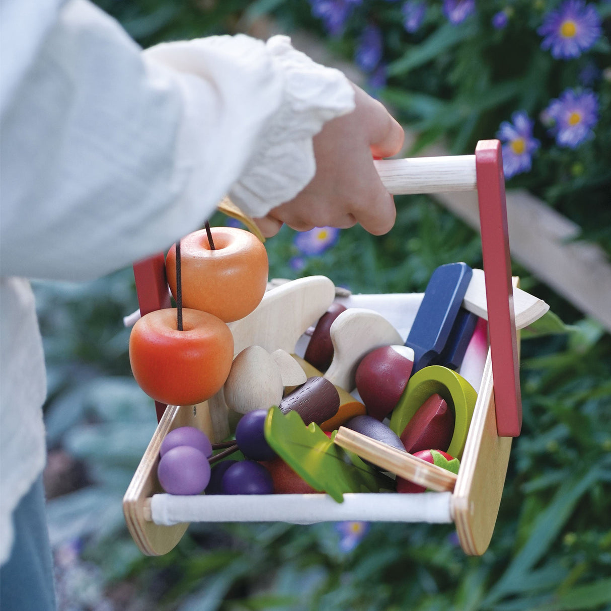 Child's hand holding a toy fruit trug with colourful wooden fruits against a natural background.