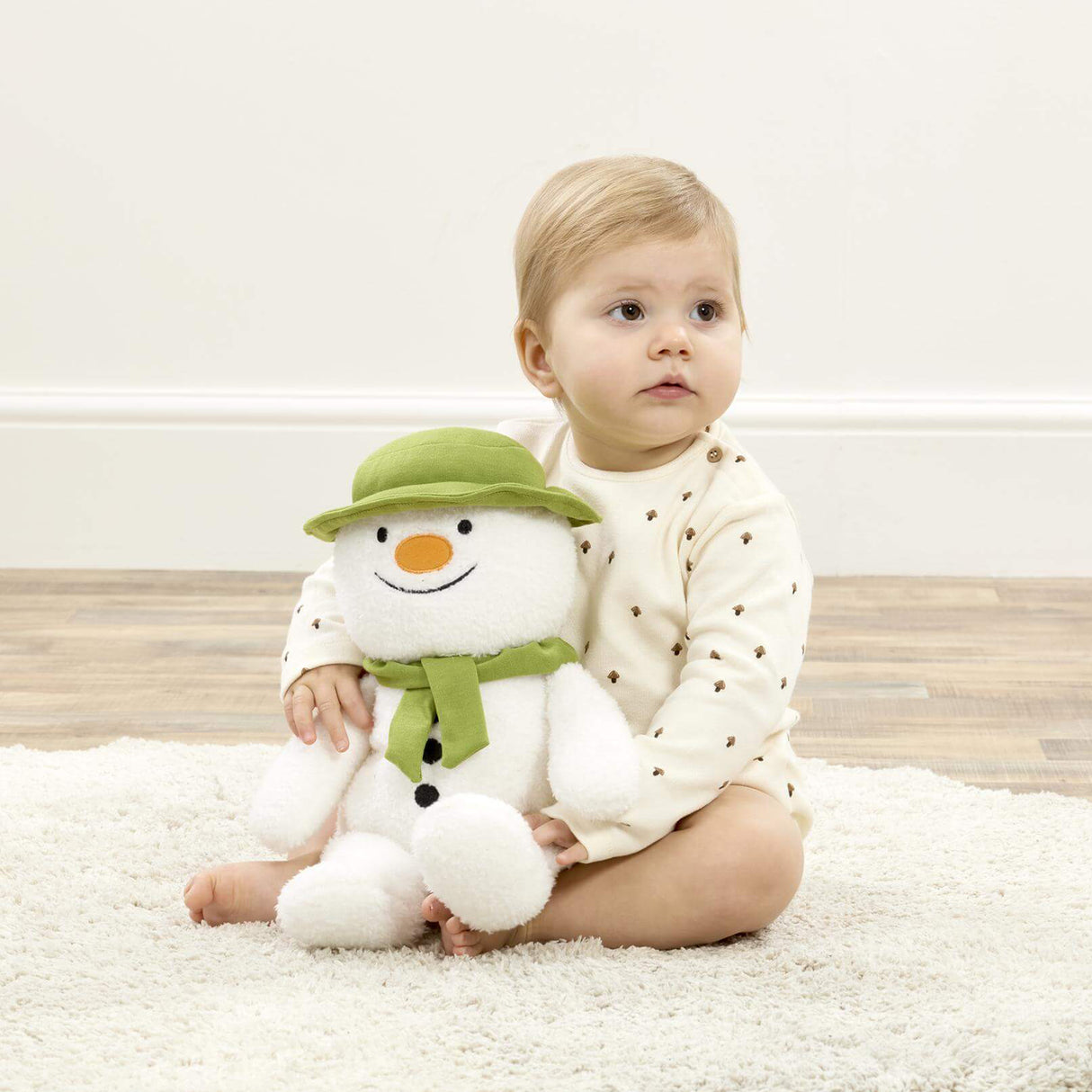 Baby holding a plush snowman toy on a light-colored rug.