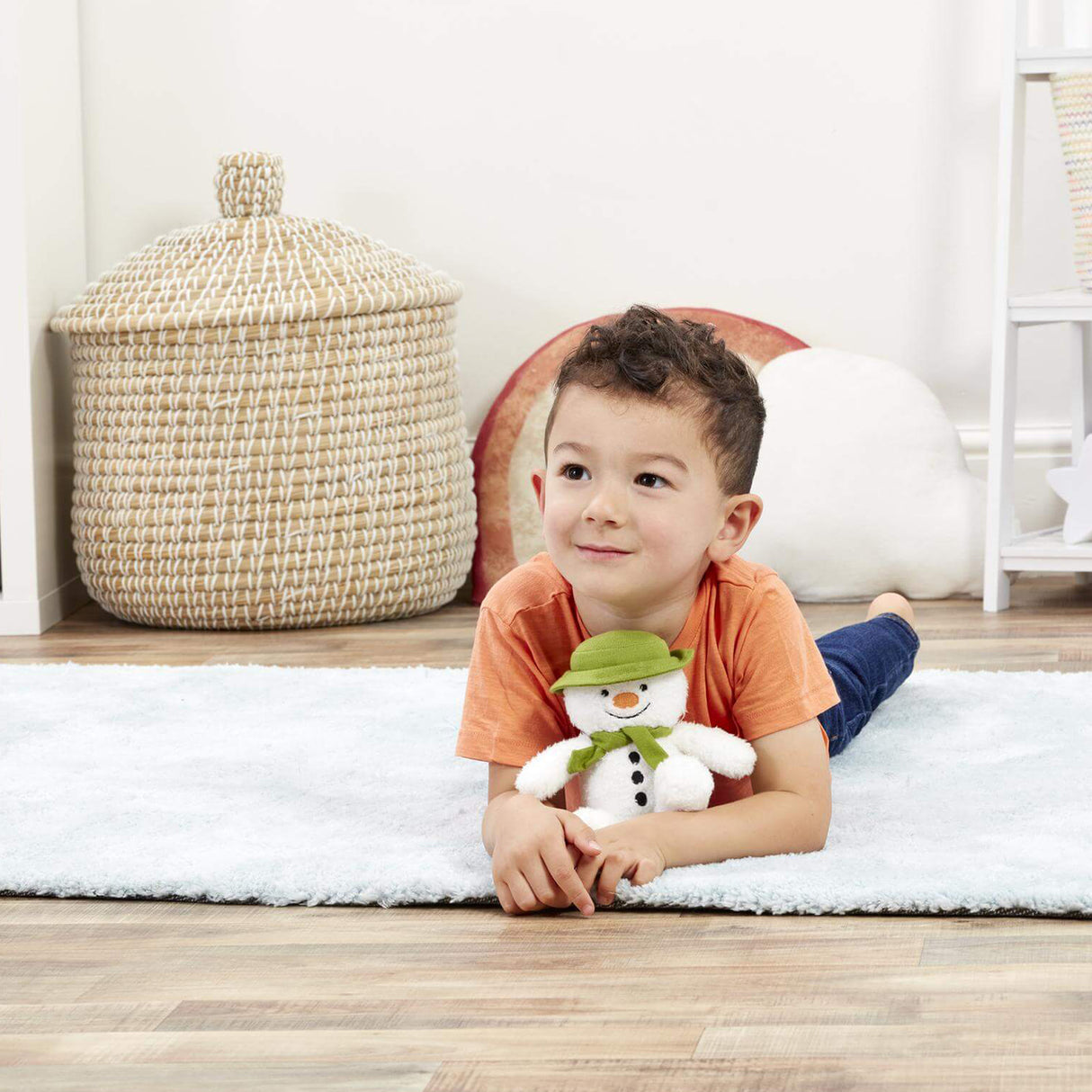 Child lying on a rug holding a snowman plush toy in a room with a woven basket and white wall.