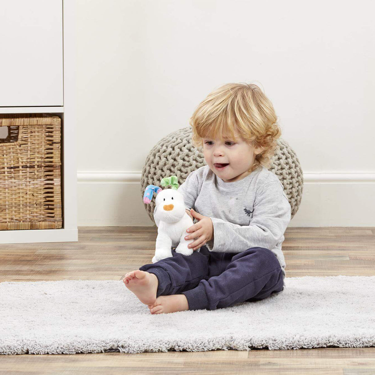 Child sitting on a rug holding a plush Snowfdog toy in a room with a wicker basket and textured chair.
