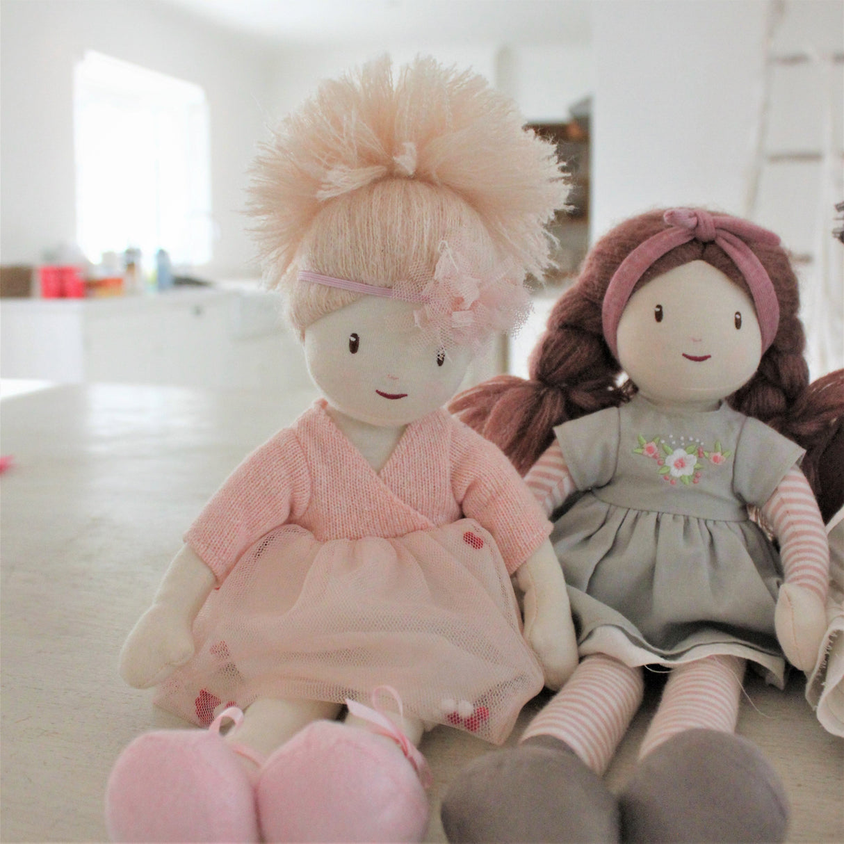 Two rag dolls, one in pink outfit and the other in gray dress with floral details, sitting on a surface.