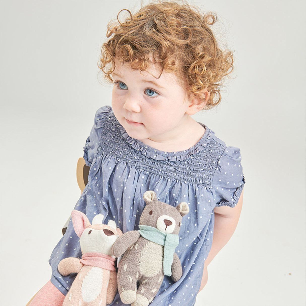 Child wearing a blue dress with polka dots, holding two stuffed animals against a light background
