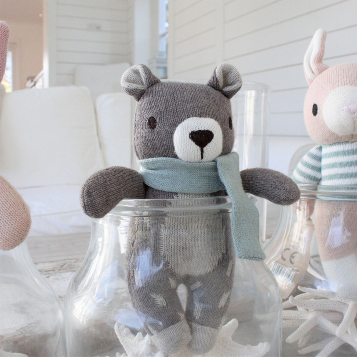 Knitted teddy bear with a light blue scarf with a blue scarf inside a glass jar, surrounded by other toys.