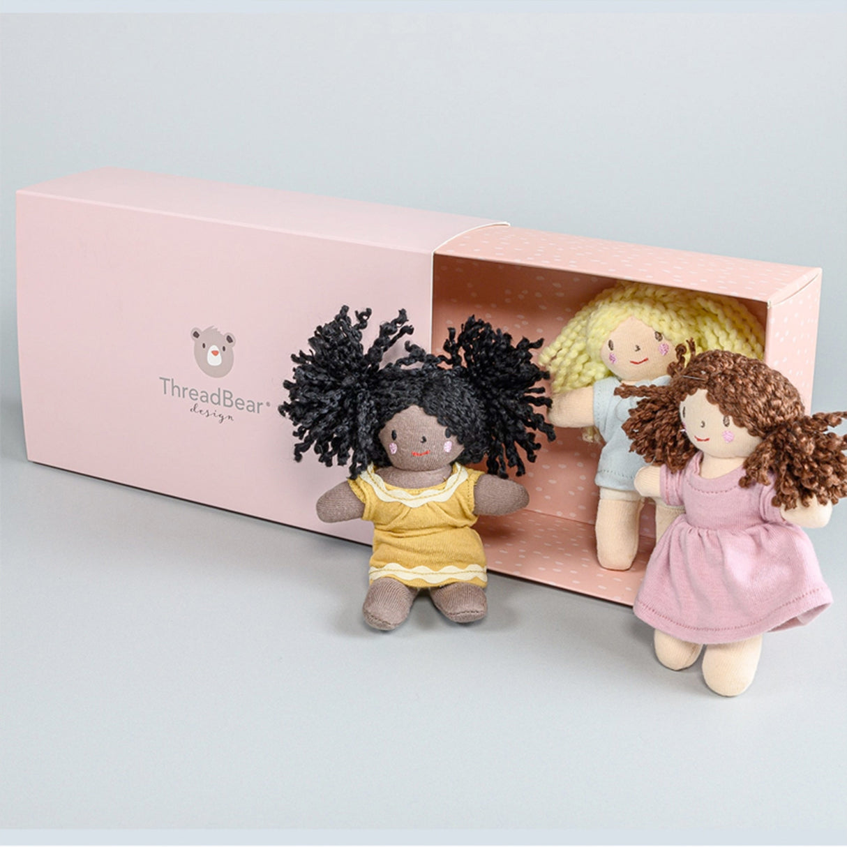 Three small rag dolls with different hairstyles and outfits in front of a pink 'ThreadBear Design' box.