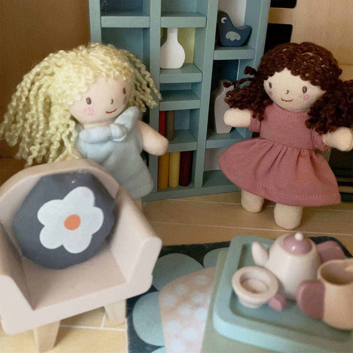 Two dolls with blonde and brown hair, a chair with a flower cushion, and a small table with a teapot set on a wooden floor.