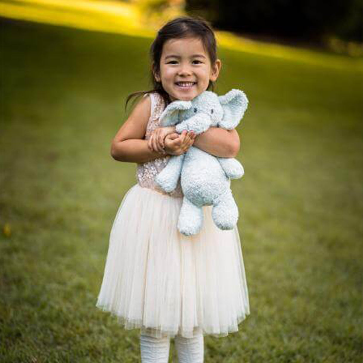 Young girl in white dress holding a stuffed organic blue elephant toy outdoors on grass