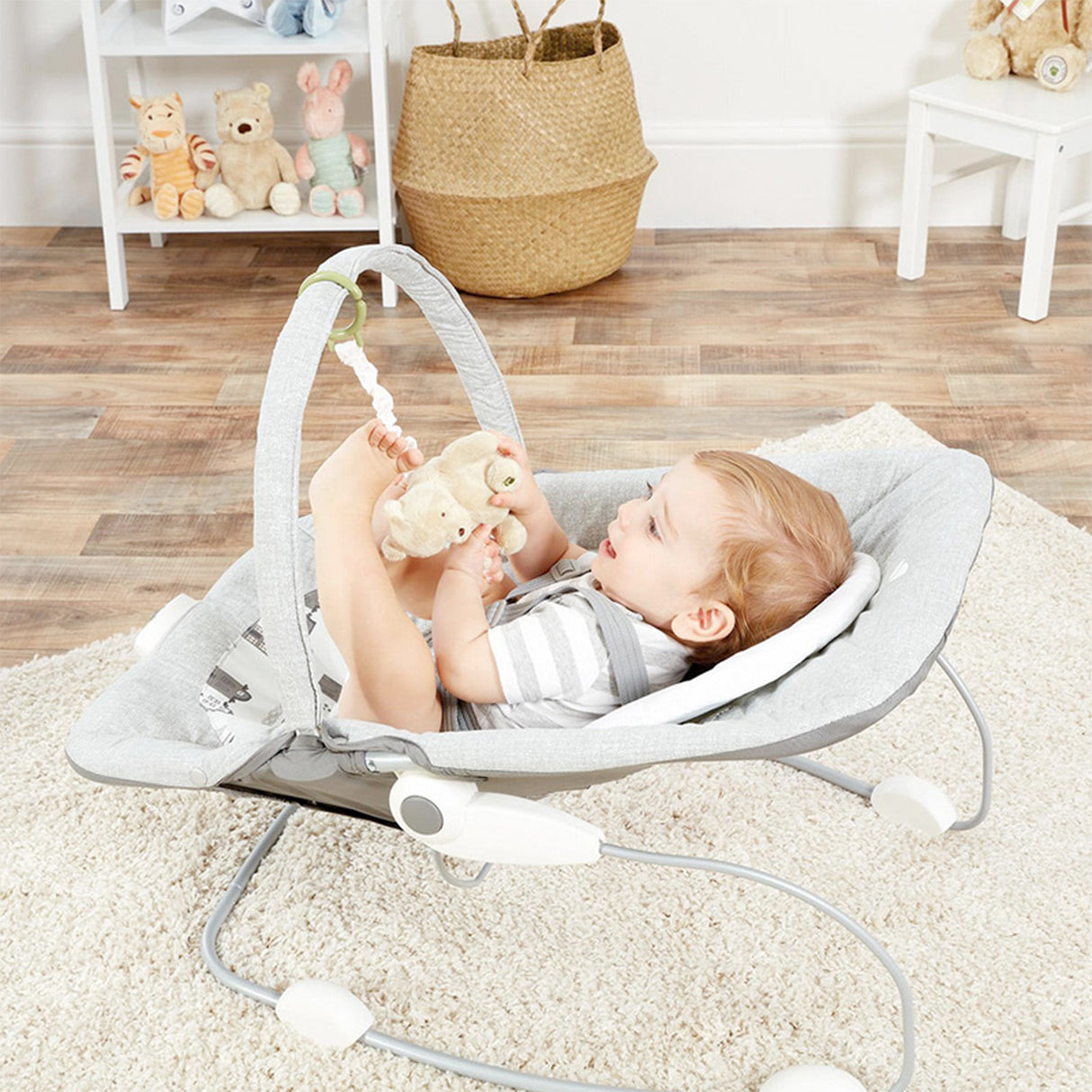 Child in a baby bouncer holding a teddy bear in a room with toys and furniture.