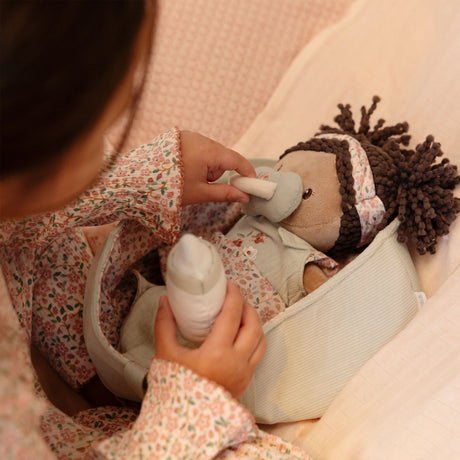 Child feeding Little Dutch Baby Doll Evi a bottle while doll lies in sage green basket