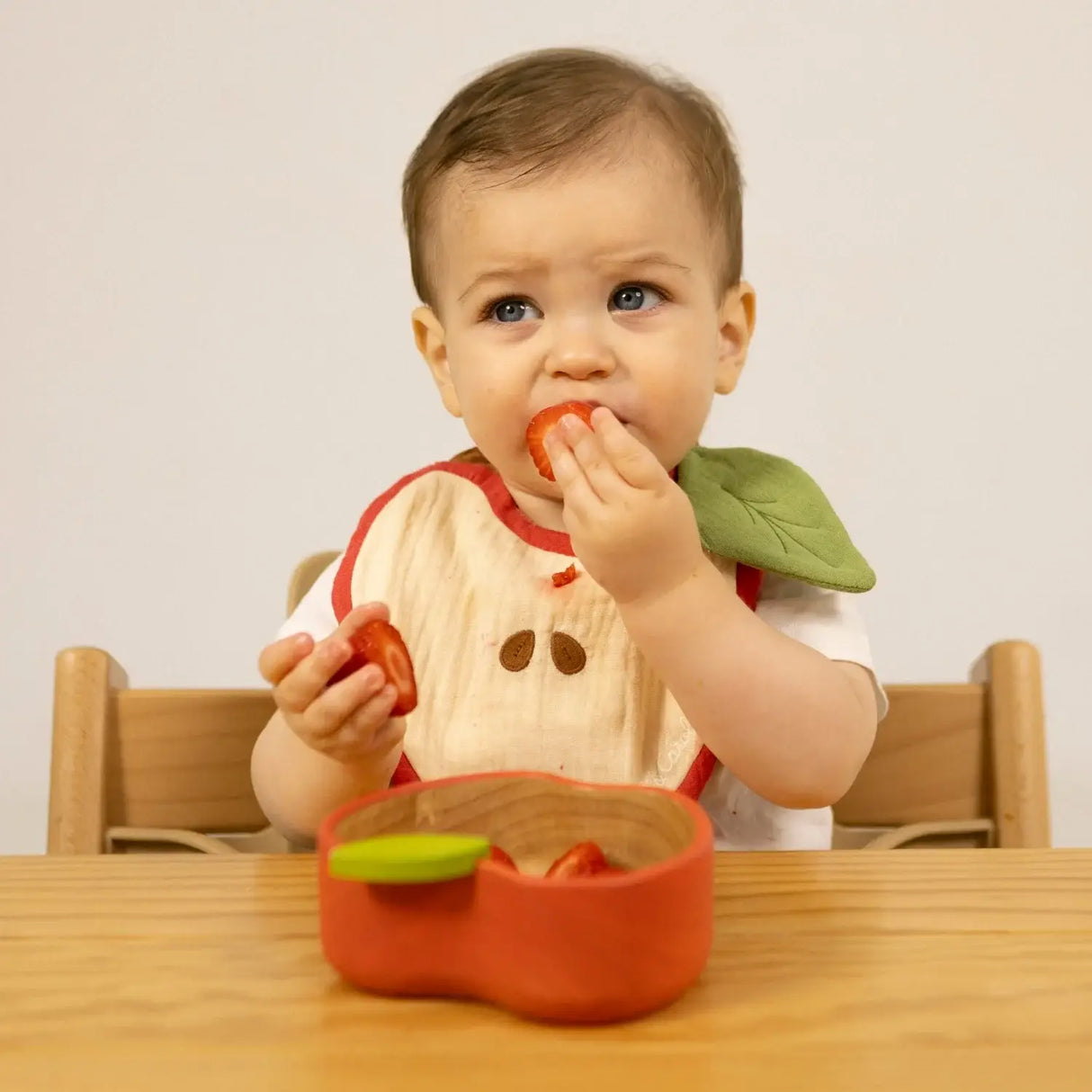 Baby eating strawberries with an apple bib at a wooden table eating from an apple shapped bowl