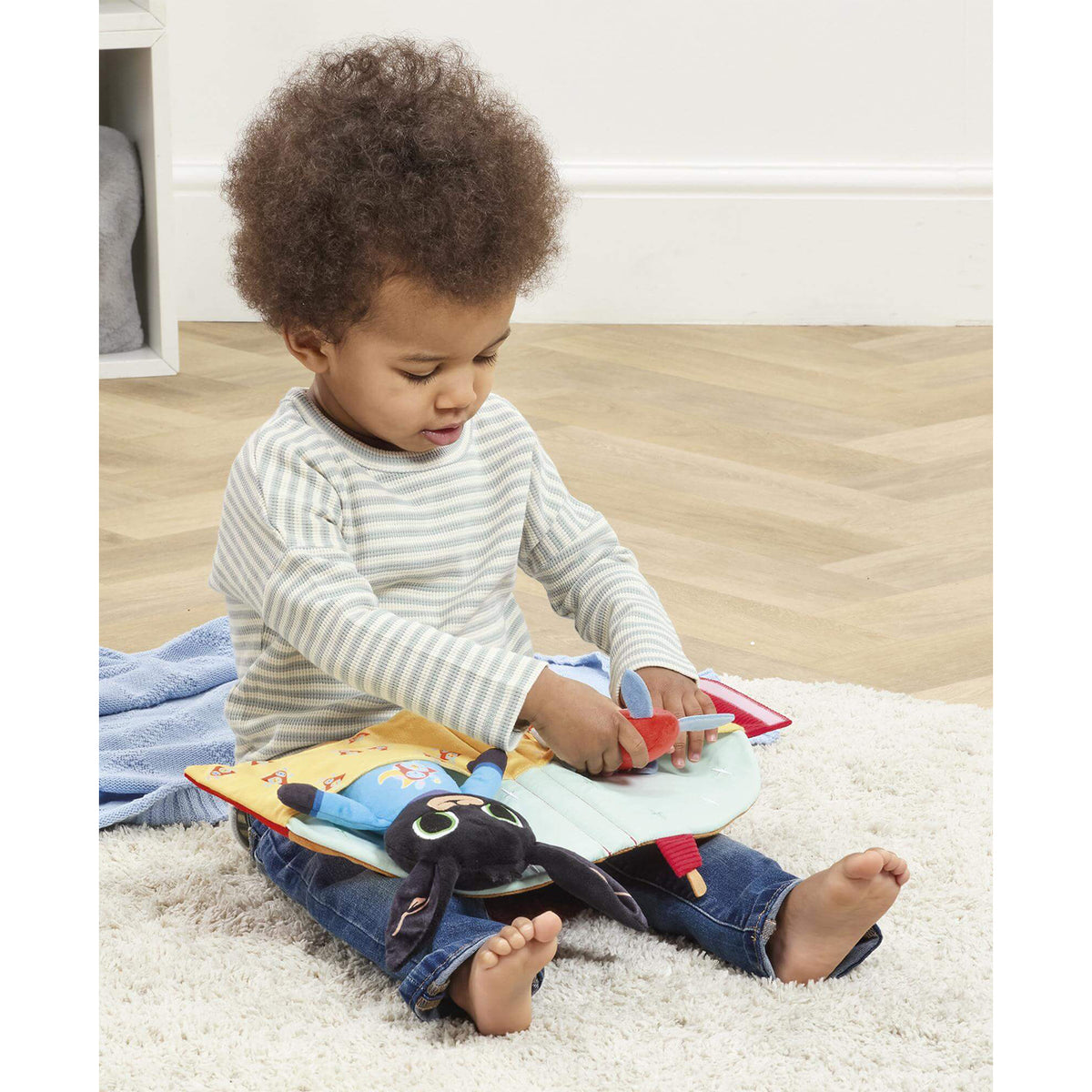 A toddler sits playing with a soft, colourful Bing travel bedtime toy.