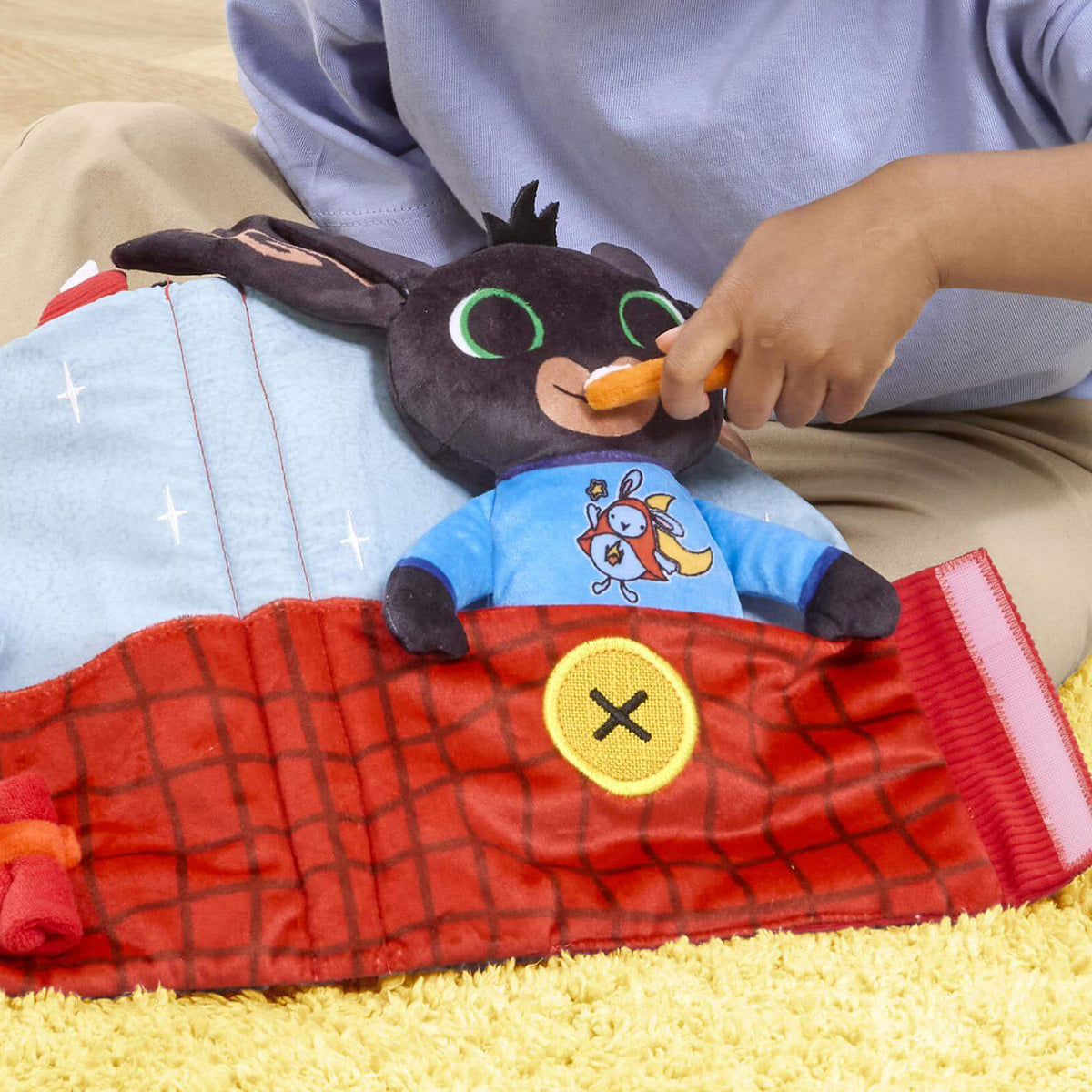 A child's hand brushes a soft toy bings teeth , bing is wearing blue pjs and red blanket with a yellow button.