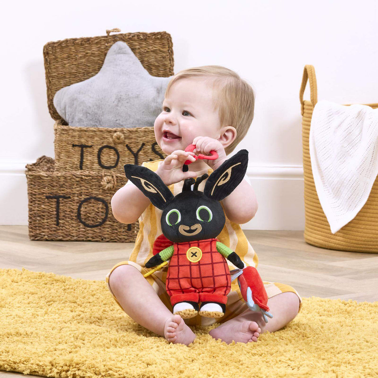 Child playing with a plush bing toy on a yellow rug, with storage baskets labeled 'TOYS' in the background.