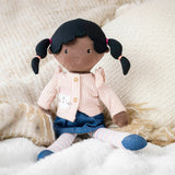 Stuffed doll with black hair in pigtails wearing a pink sweater and blue dress on a soft surface.