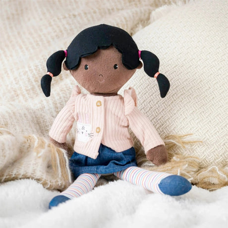 Stuffed doll with black hair in pigtails wearing a pink sweater and blue dress on a soft surface.