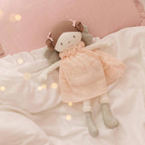 Rag doll with a pink muslin dress lying on a soft surface with blurred lights in the background
