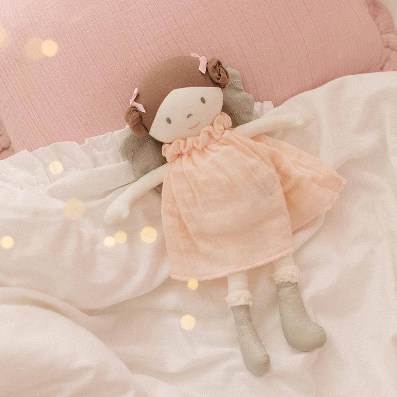 Rag doll with a pink muslin dress lying on a soft surface with blurred lights in the background
