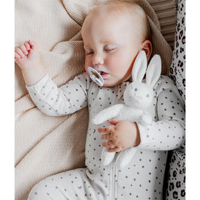 Baby sleeping with a soft bunny toy and dummy, wearing a polka dot onesie.
