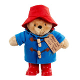 Stuffed Paddington toy bear in a blue coat, red hat, and boots with a tag on a white background