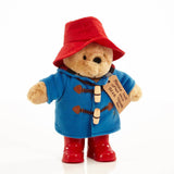 Stuffed classic Paddington Bear soft toy with red hat, blue coat with wooden toogles, red boots and a tag 'Please look after this bear' on a white background.