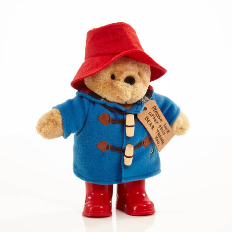 Stuffed classic Paddington Bear soft toy with red hat, blue coat with wooden toogles, red boots and a tag 'Please look after this bear' on a white background.