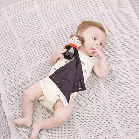 Baby lying on a textured blanket wearing a onesie holding a Harry Potter themed comfort blanket with a plush toy of Harry Potter in his signature school robes and Gryffindor scarf attached to the corner.