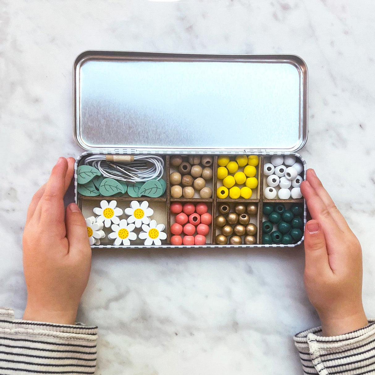 Open tin with compartments filled with colourful beads and decorative items, held by two hands on a marble surface.