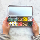 Open tin with compartments filled with colourful beads and decorative items, held by two hands on a marble surface.
