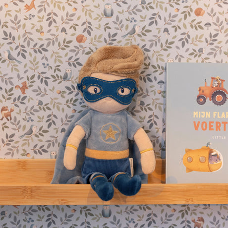 Stuffed toy in superhero costume with a book on a shelf against a patterned wall.