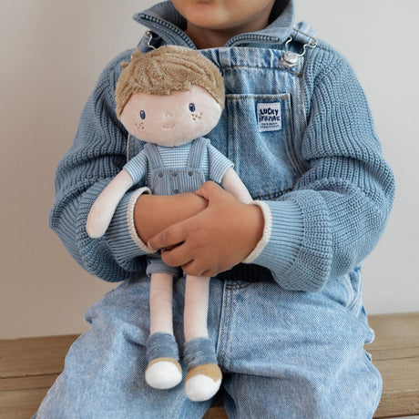 Child holding a soft boy doll in blue overalls and a stripey blue t-shirt, blue socks and brown shoes.