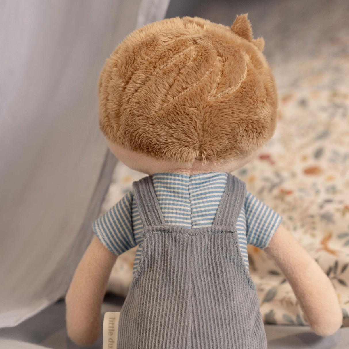 back view of a male soft doll with blonde hair, blue overalls and a stripey blue t-shirt.