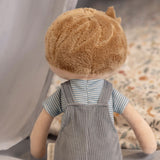 back view of a male soft doll with blonde hair, blue overalls and a stripey blue t-shirt.