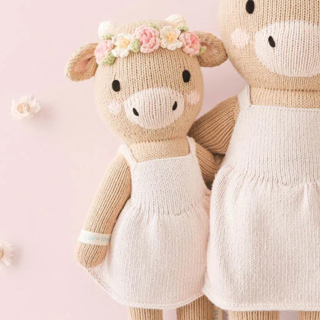 Knitted soft toy cows with a pale pink dresses and floral pink and cream headband on a pink background