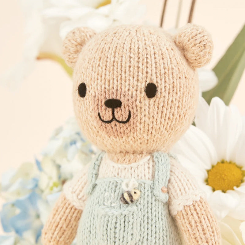 Knitted teddy bear with a light blue dungarees surrounded by flowers